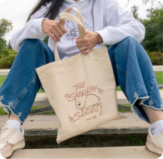 Canvas tote bag held by a seated model, printed with the Scholars Society bear artwork