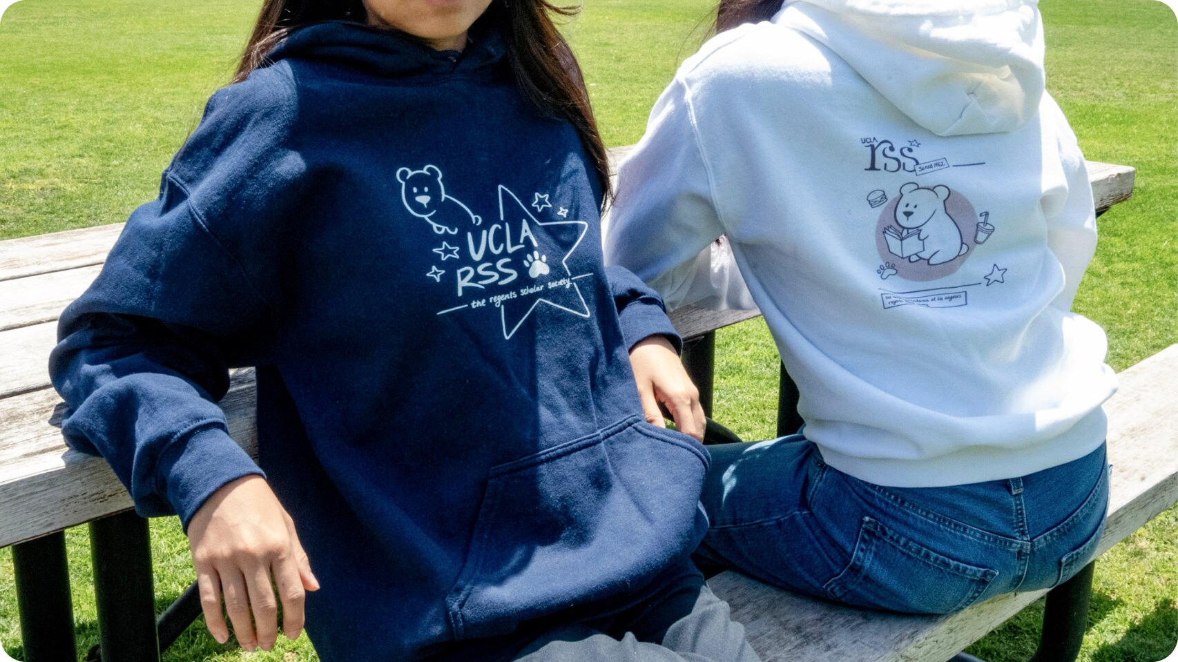Two students on a bench wearing the RSS hoodies — the navy front with the UCLA RSS star, and the white back with the bear design