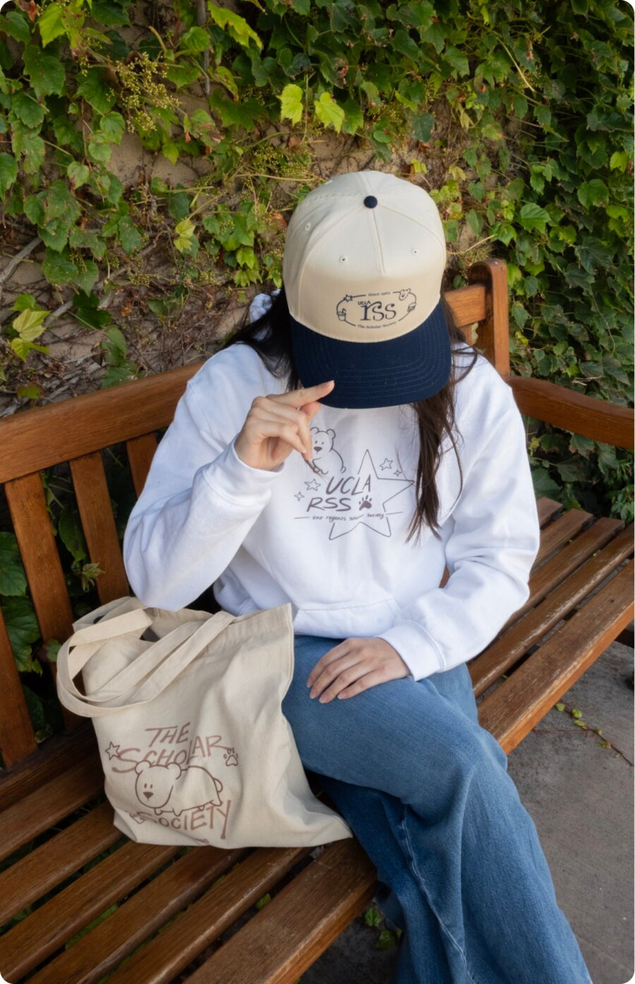 A model in the white hoodie seated on a wooden bench, holding the canvas tote