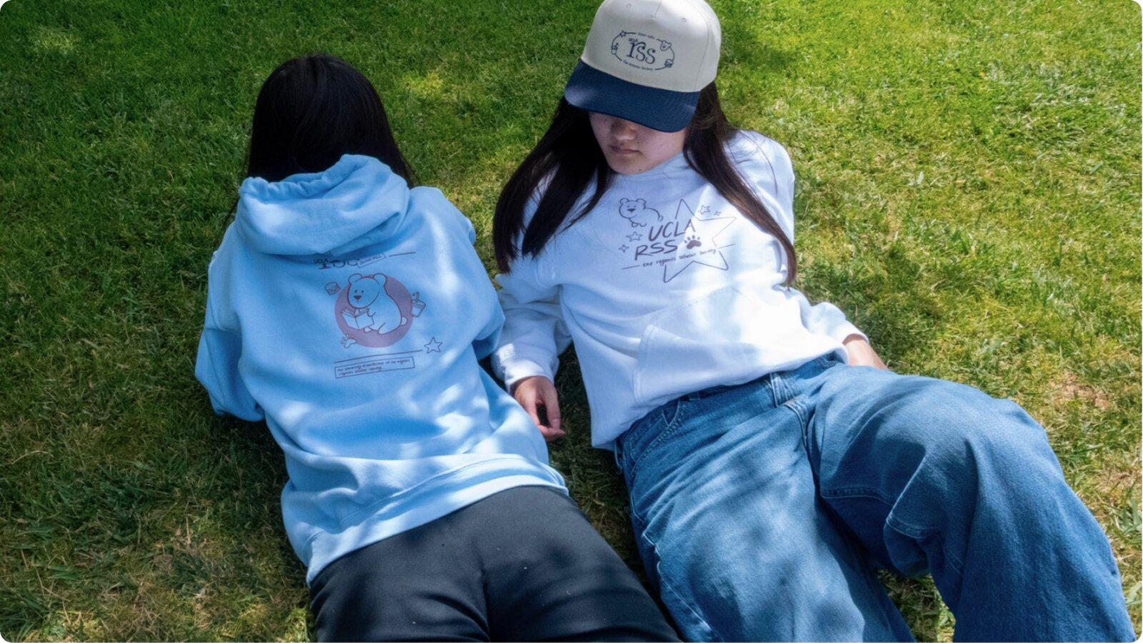 Two models lying on the grass in the light-blue and white hoodies