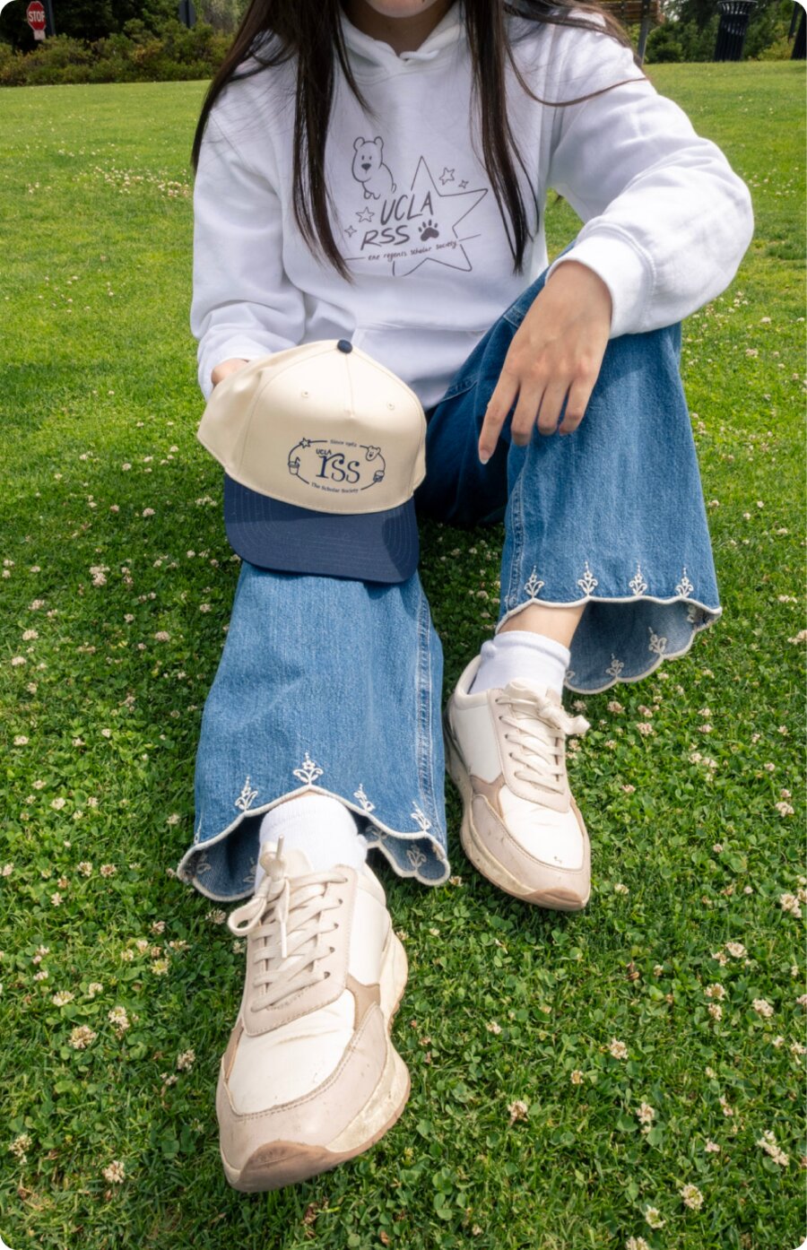 A model seated on the grass with the cap resting on one knee and cream sneakers