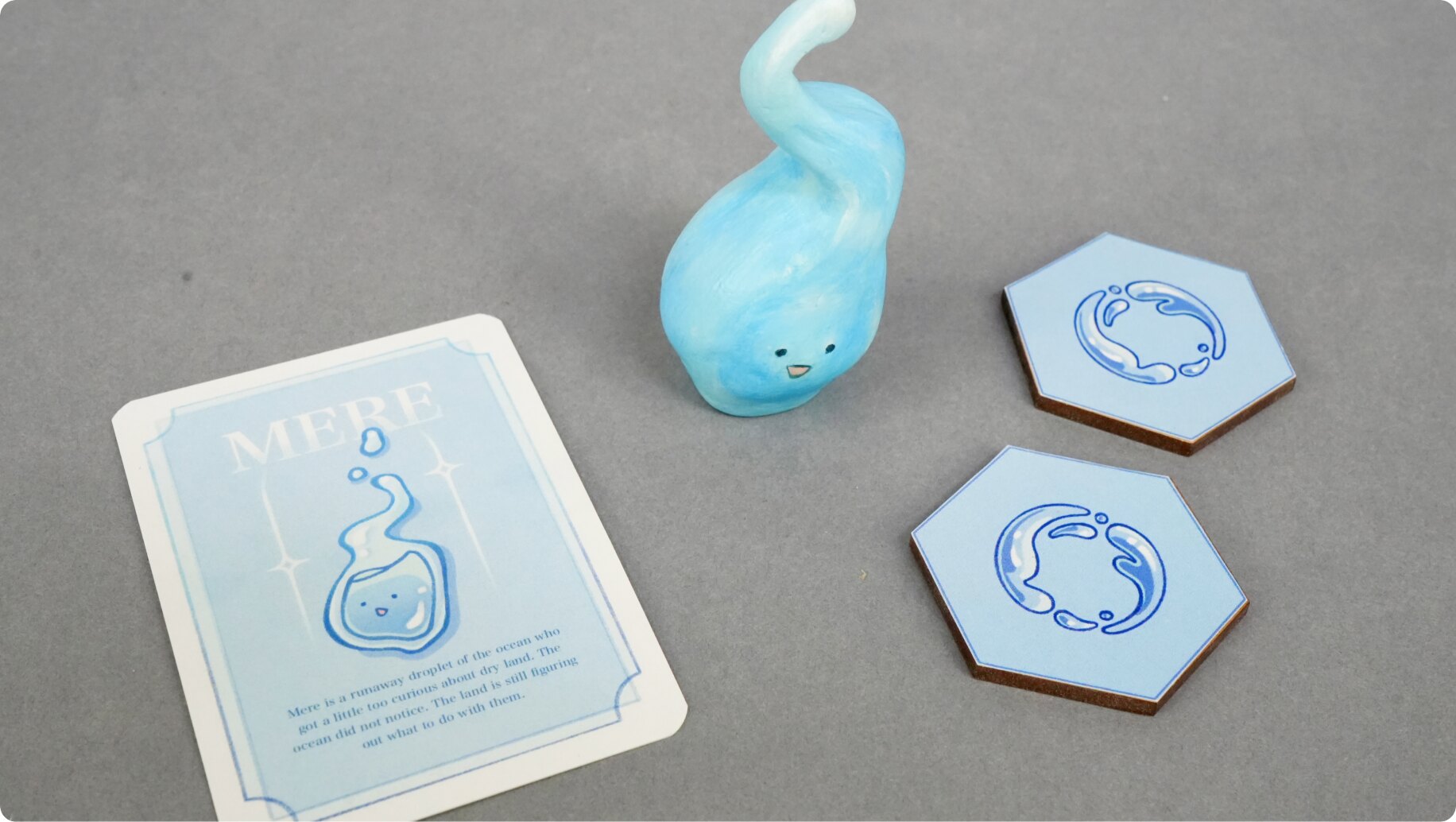 Mere's character card, clay miniature, and two water hex tiles arranged together